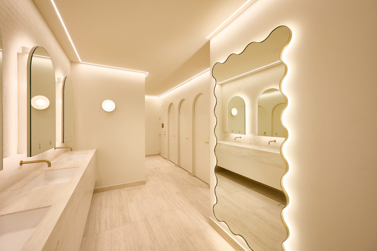 An amenities corridor with a tall scalloped backlit mirror and a row of arched backlit mirrors above a stone vanity
